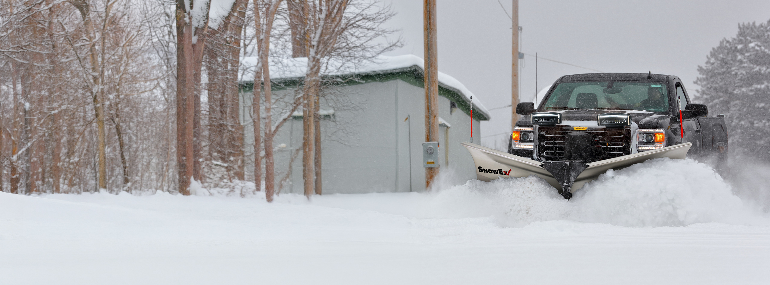 A black pickup truck with a large snowplow attachment clears deep snow from a road beside a building on a snowy, overcast day. The powerful plow pushes snow forward as the truck steadily advances.