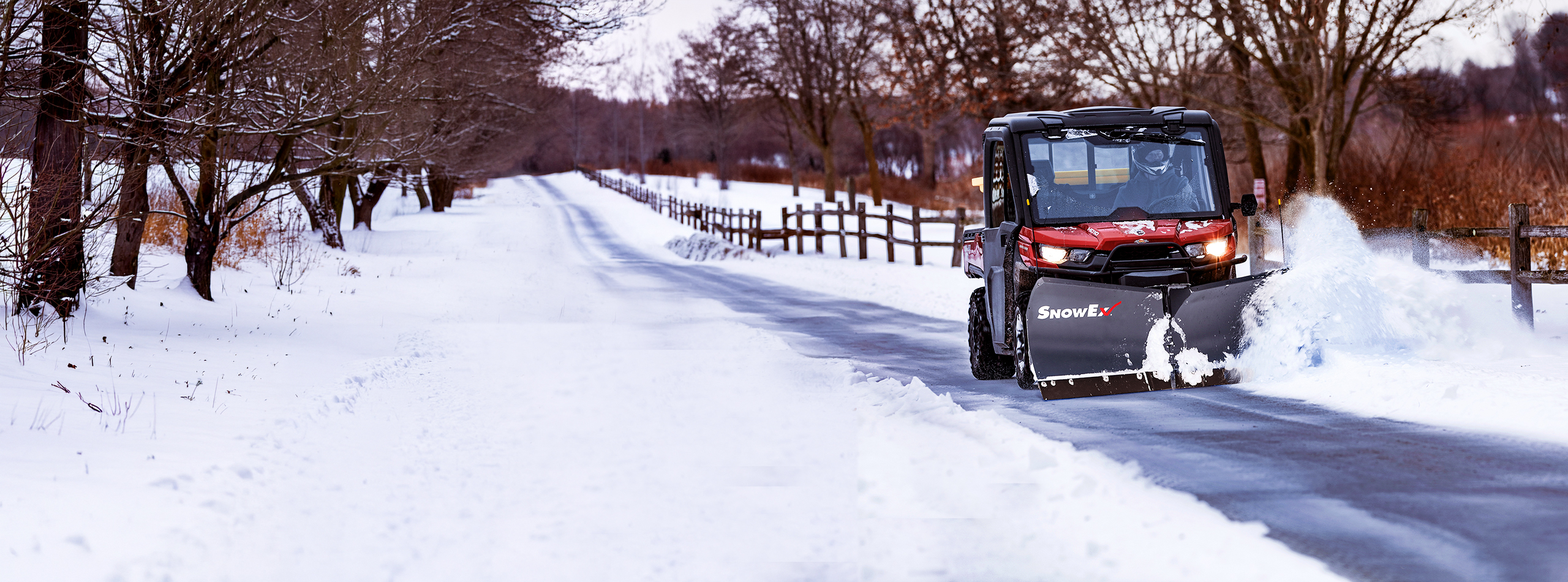 A utility vehicle with a SnowEx plow attachment clears snow from a road on a crisp winter day, with trees and a fence in the background.
