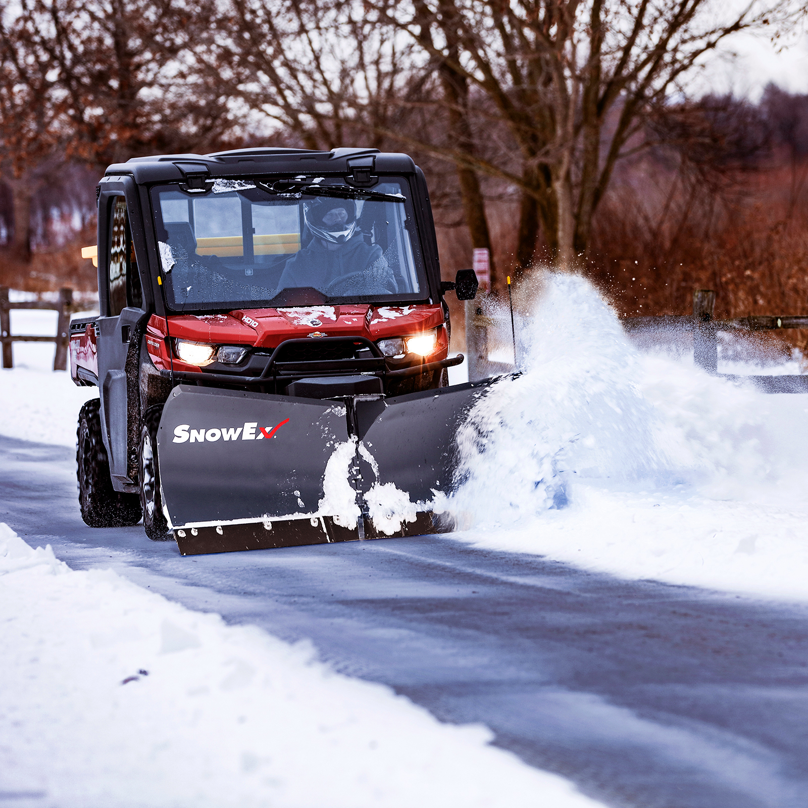 A utility vehicle with a SnowEx plow attachment clears snow from a road on a crisp winter day, with trees and a fence in the background.