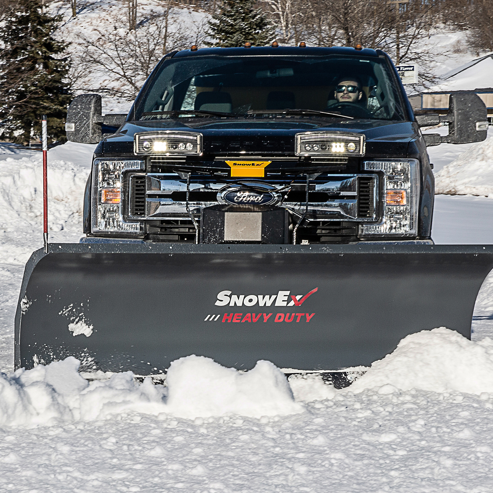 A black truck with a SnowEx heavy-duty plow clears snow from a parking lot on a sunny winter day, with snow-covered trees and buildings in the background.