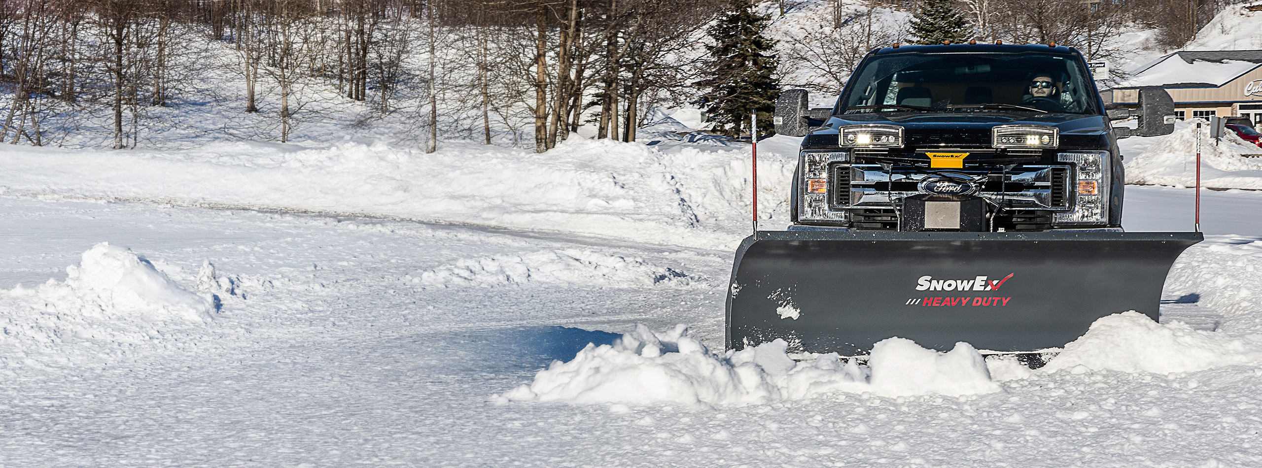 A black truck with a SnowEx heavy-duty plow clears snow from a parking lot on a sunny winter day, with snow-covered trees and buildings in the background.