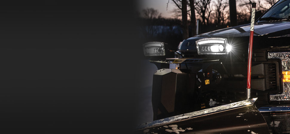 A close-up of the front of a black pickup truck with headlights on, fitted with a snow plow blade, parked outdoors at dusk with trees in the background.