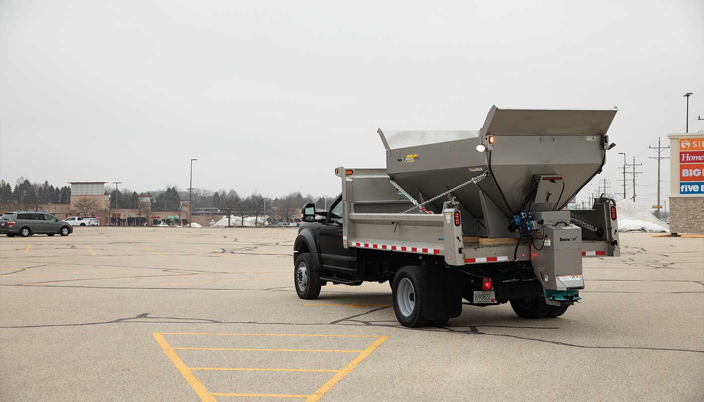 A black dump truck with a metal salt spreader mounted on its bed is parked in an empty shopping center lot on a cloudy day. Store signs and a few cars are visible in the background, highlighting the truck's utility in winter maintenance.