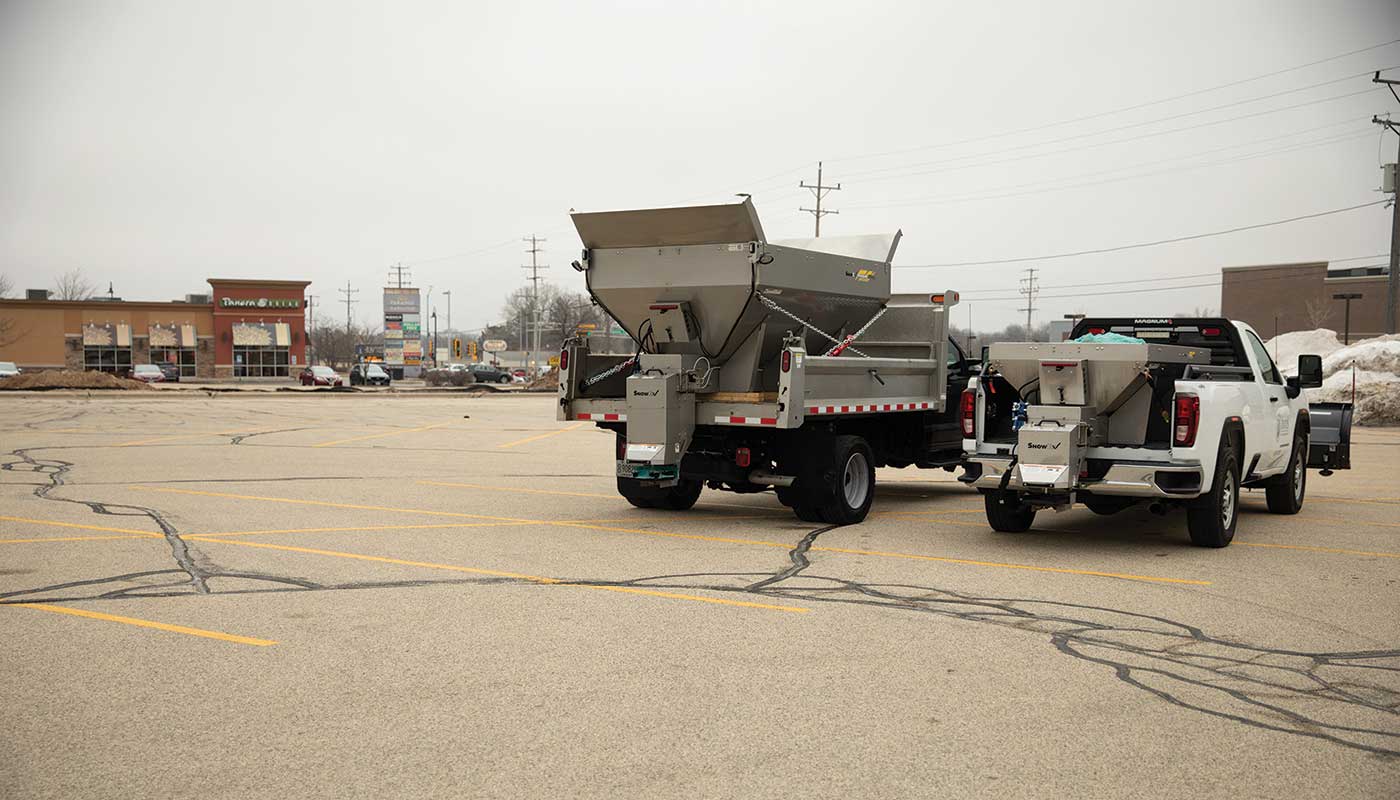 Two trucks with large salt spreaders are parked in an empty, cracked parking lot on a cloudy day, with buildings and snow piles visible in the background.