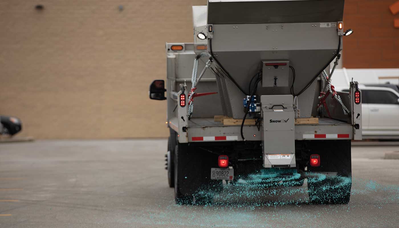 A salt spreader truck disperses blue road salt onto a parking lot, with the salt visibly scattering behind the vehicle. The truck is parked near a tan brick building, efficiently improving winter safety.