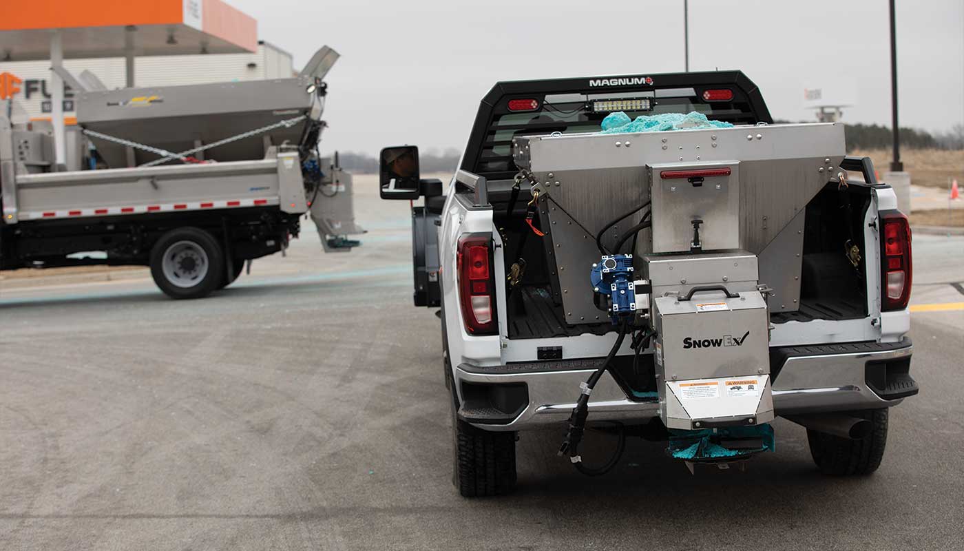 A white pickup truck with a salt spreader attached to its bed is parked near a similar truck in a parking lot on an overcast day. An industrial building and an orange gas station canopy are visible in the background, emphasizing utility and reliability.