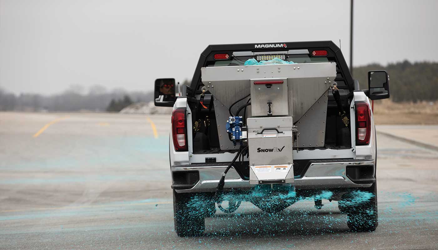 A pickup truck equipped with a large salt spreader disperses granular blue de-icing material on an empty, overcast road, preparing the surface for icy conditions.