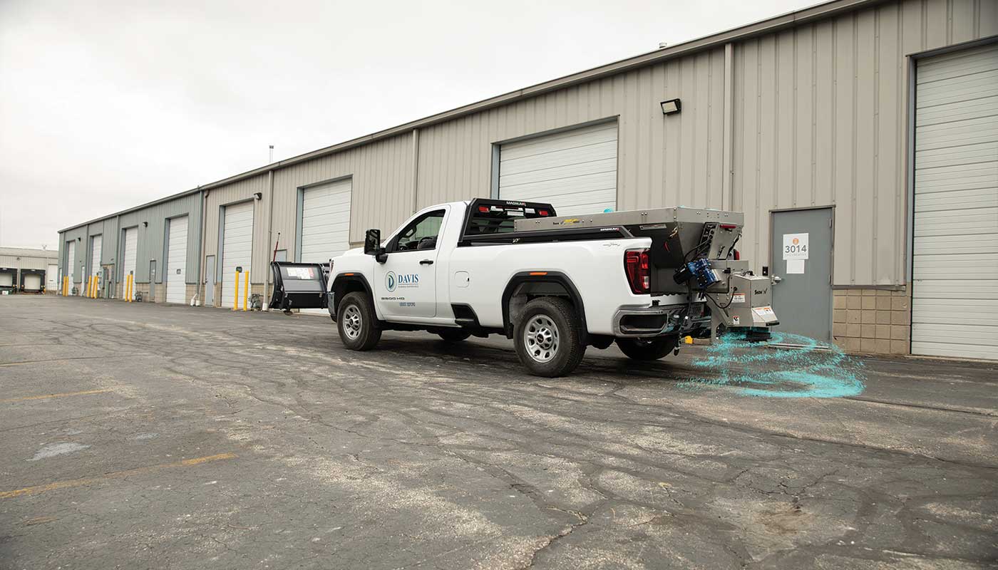 A white pickup truck with a salt spreader is distributing blue de-icing material on the pavement outside a row of industrial garage doors, helping keep surfaces safe during winter conditions.