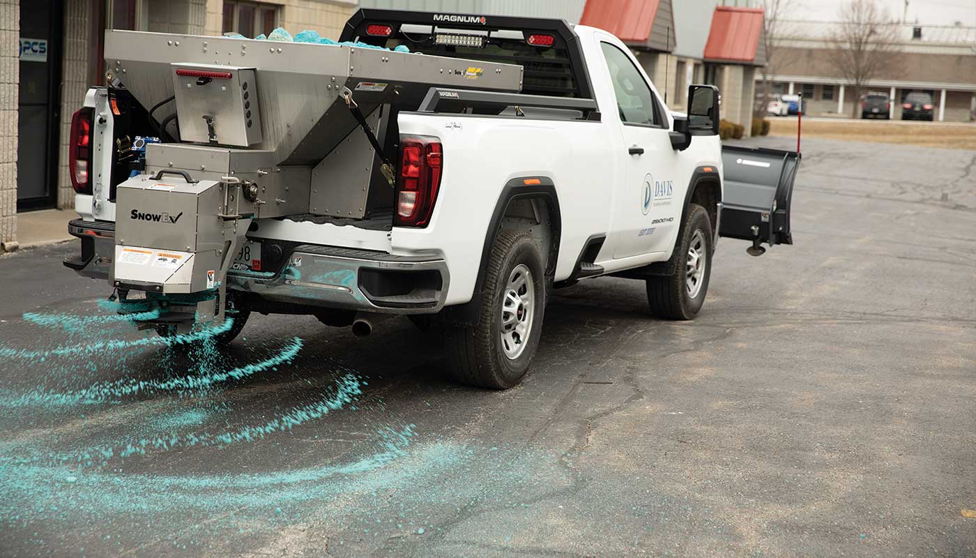 A white pickup truck spreads blue de-icer onto a paved parking lot using equipment mounted on its back, preventing ice buildup near commercial buildings.