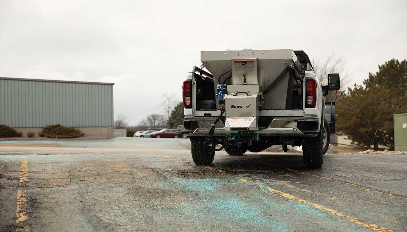 A pickup truck with a large salt spreader attached to the bed is distributing blue de-icer on a parking lot, with buildings and trees visible in the background.