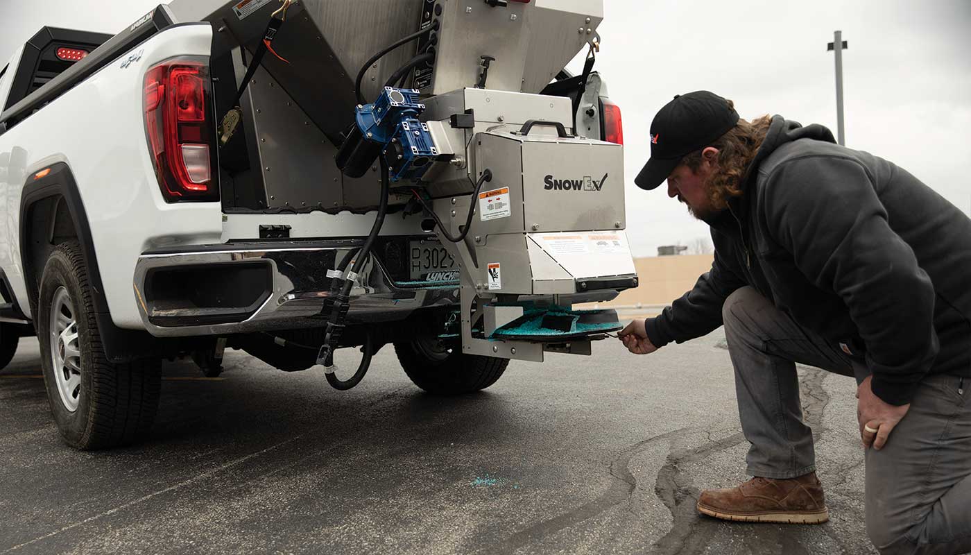 A person kneels beside a white pickup truck, checking a SnowEx salt spreader attached to the back in a parking lot under an overcast sky.