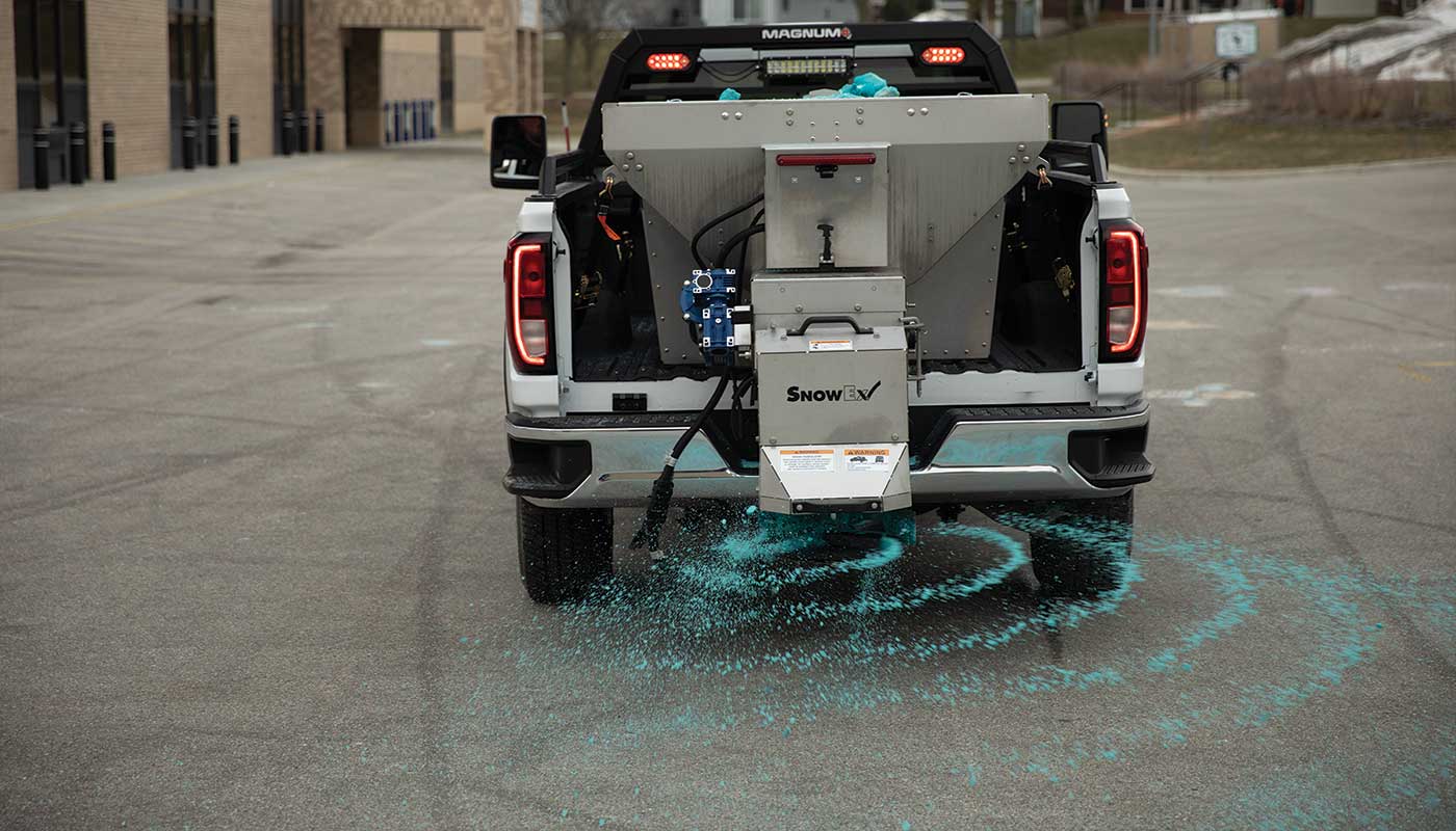 A truck with a mounted spreader dispenses blue de-icing material onto an asphalt surface in a parking lot, creating a circular pattern of sodium-based material behind the vehicle.