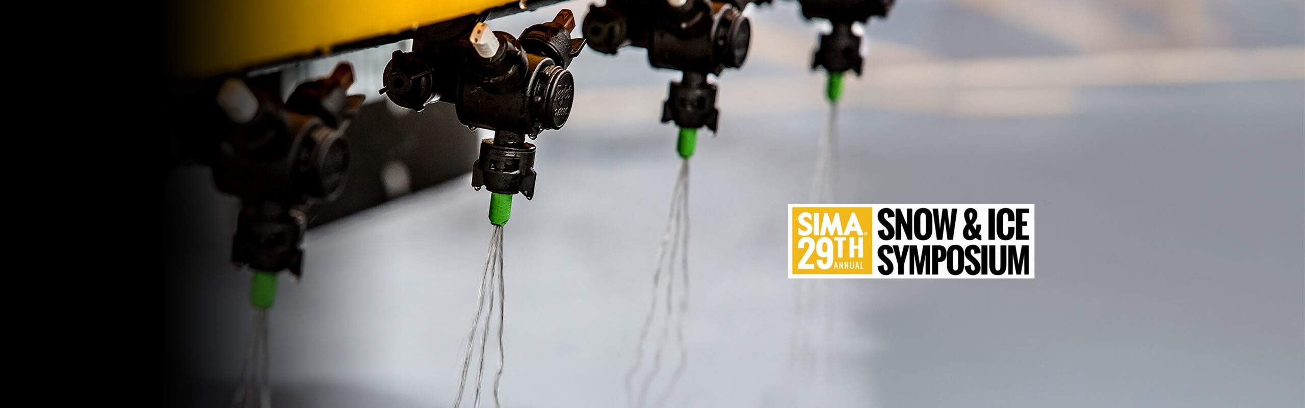 Close-up of several spray nozzles releasing streams of liquid, with a blurred background. Text overlay reads “SIMA 29th Annual Snow & Ice Symposium.”.