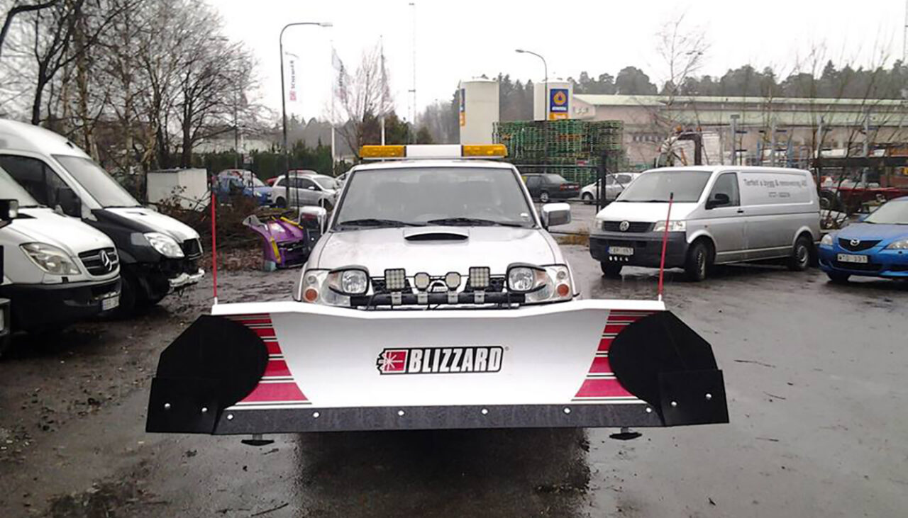 A silver pickup truck with a large Blizzard snow plow attached to the front is parked in a lot with other vehicles on a wet, overcast day.
