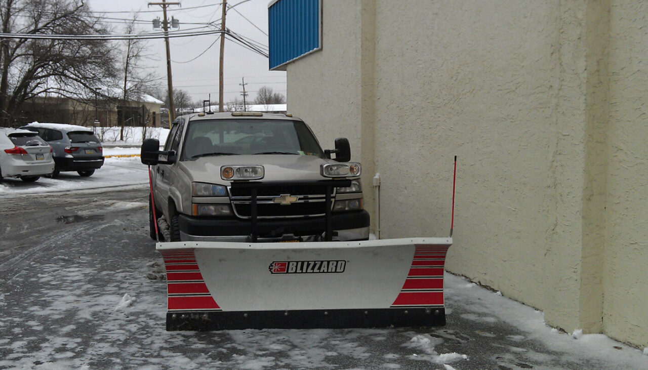 A snow plow truck with Blizzard branding is parked on a snowy and icy lot next to a beige building, with other vehicles and trees visible in the background.