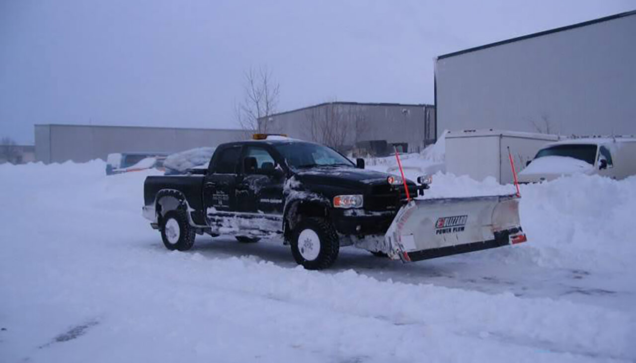 A black pickup truck with a large snow plow attached to the front clears snow from a parking lot, surrounded by deep snowbanks and industrial buildings in the background.