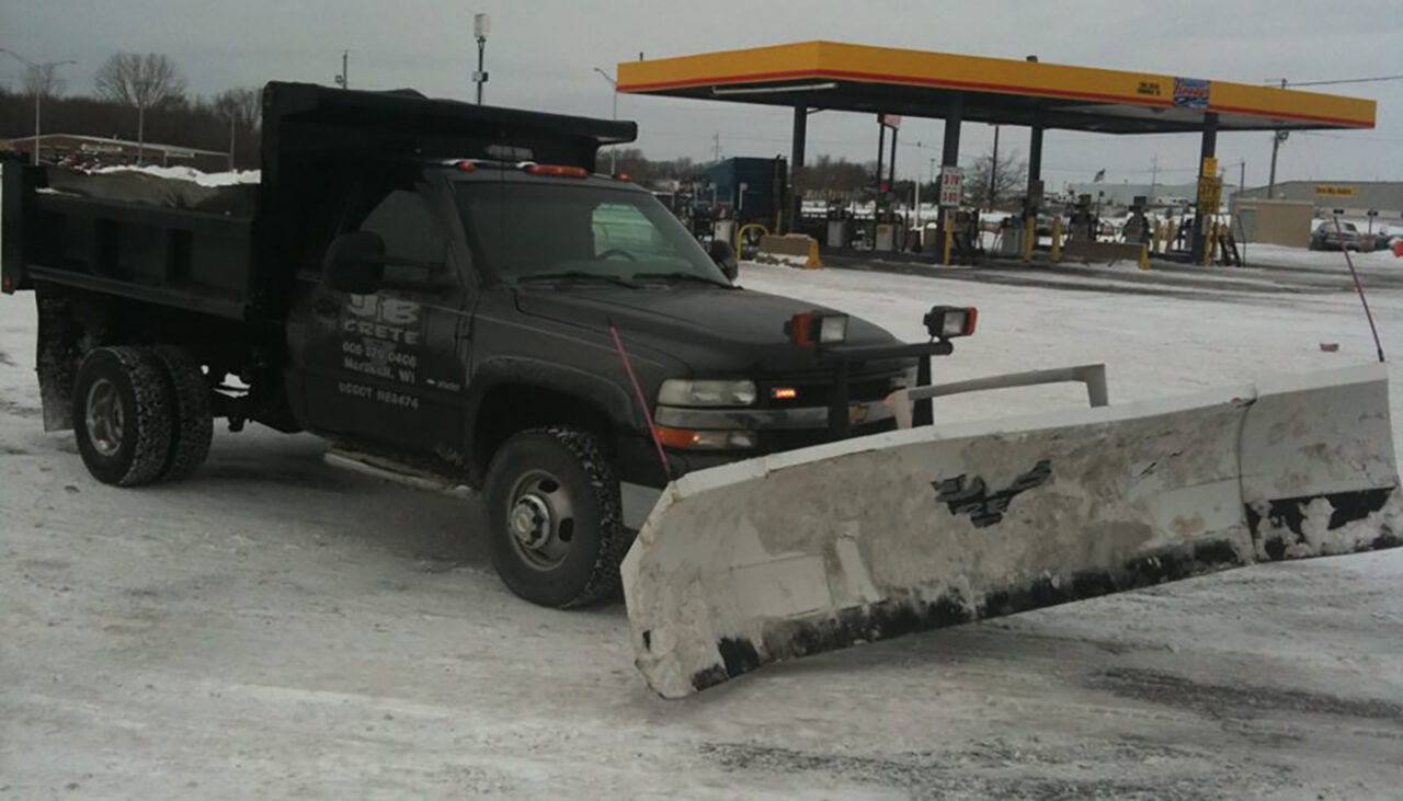 A black dump truck with a large snow plow attached to the front is parked on a snowy lot near a gas station. Snow and ice are visible on the ground and the trucks plow blade.