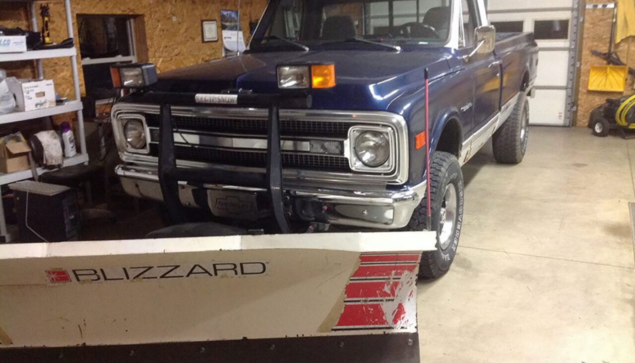 A blue vintage pickup truck with a large white Blizzard snow plow attached to the front is parked inside a garage or workshop. Shelves and tools are visible along the walls.