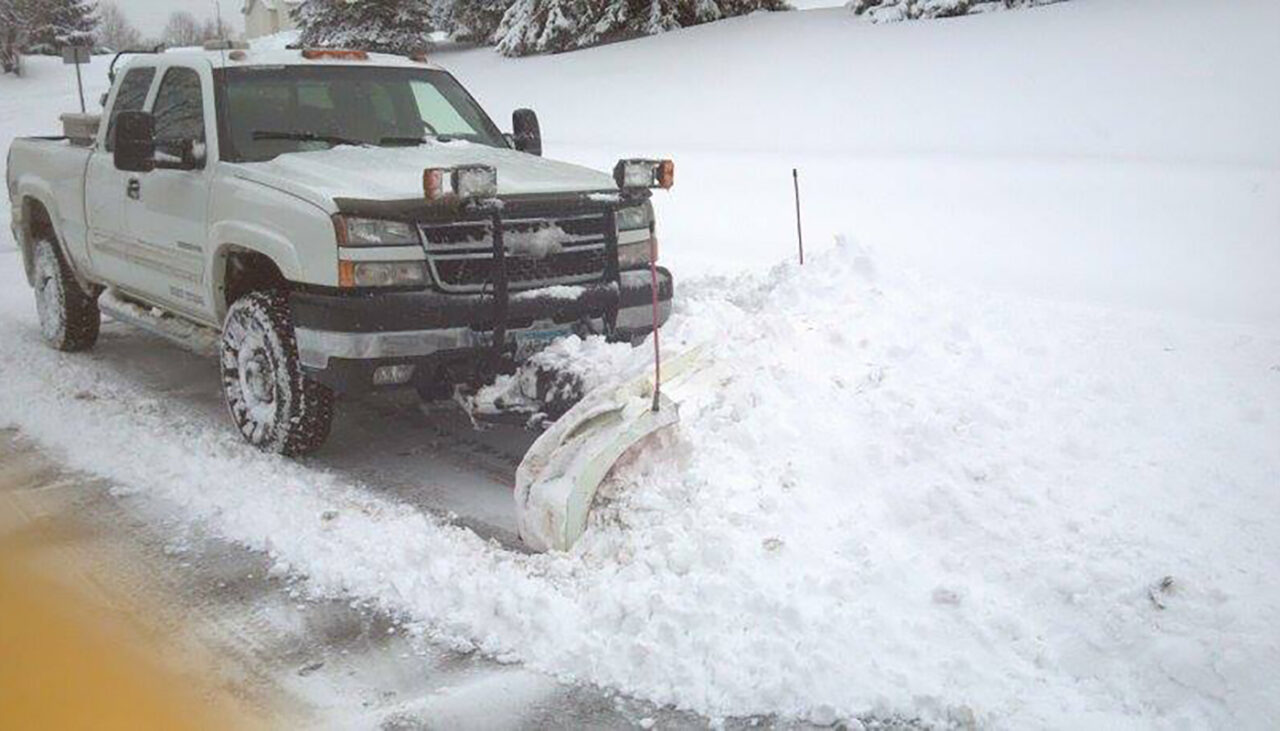 A white pickup truck fitted with a snow plow is clearing snow from a snow-covered road, pushing a large pile of snow to the side. Snow blankets the ground and trees in the background.