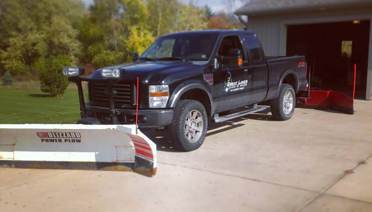 A black pickup truck equipped with a Blizzard Power Plow snow plow attachment is parked on a concrete driveway near a garage, with green grass and trees in the background.