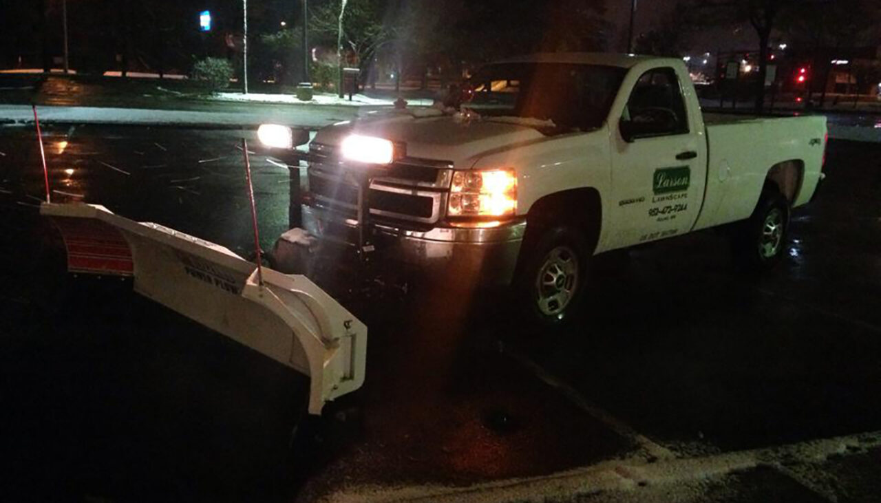 A white pickup truck with a snow plow attachment is parked in a dark, wet parking lot at night with its headlights shining. The truck has a business logo on the door. Snow is lightly falling.