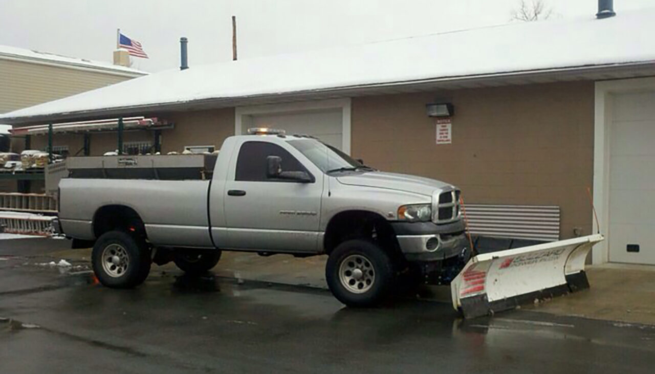 A silver pickup truck equipped with a snow plow attachment is parked on wet pavement in front of a building with a snow-covered roof. An American flag is visible in the background.
