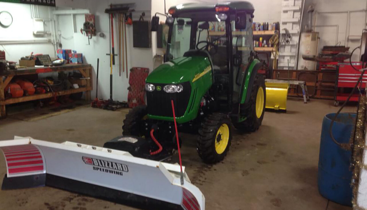 A green tractor with yellow wheels is parked indoors in a workshop. Attached to the front is a Blizzard Speedwing snowplow blade. Shelves, tools, and equipment are visible in the background.