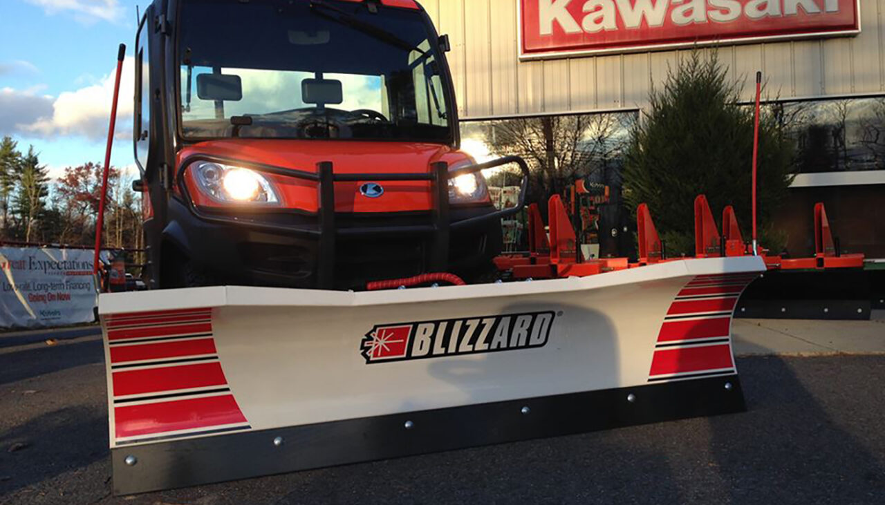 A red utility vehicle with headlights on is equipped with a large Blizzard snow plow blade, parked in front of a building with a Kawasaki sign.