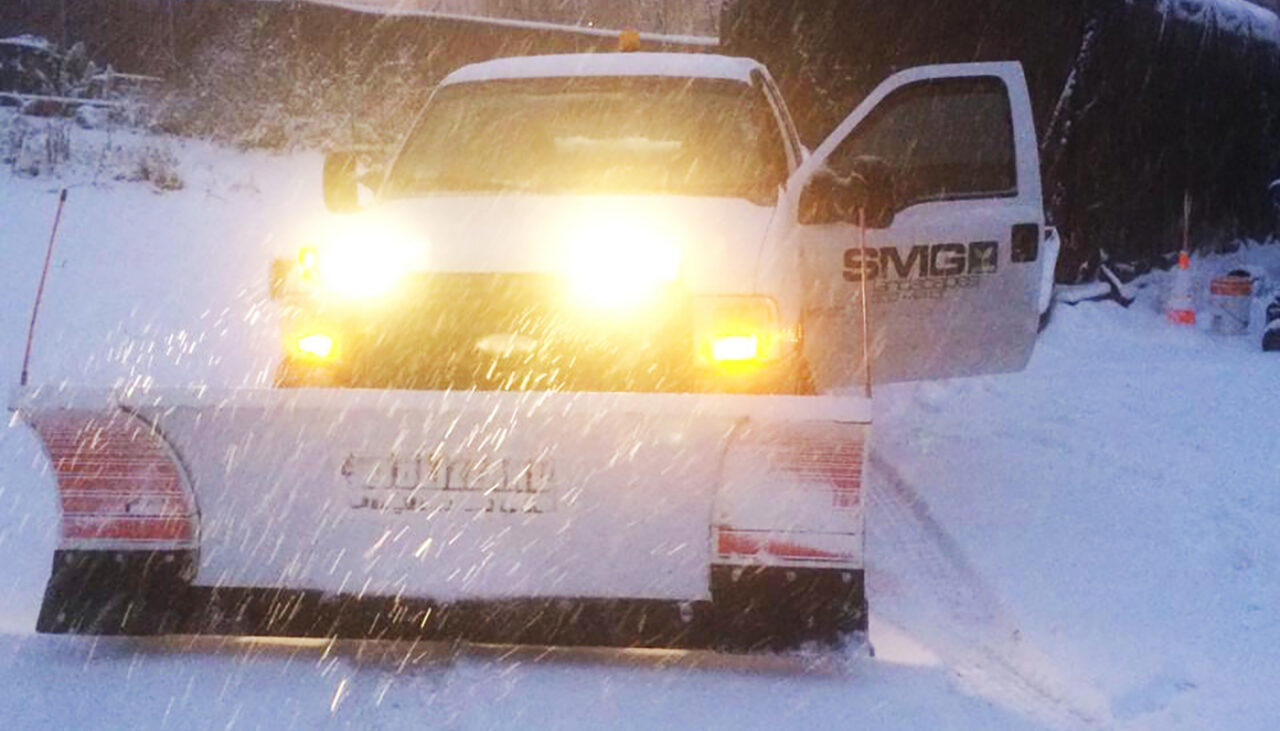 A white snow plow truck with headlights on is clearing snow in a snowy area. The drivers side door is open, and snow is falling heavily, partially obscuring the view.