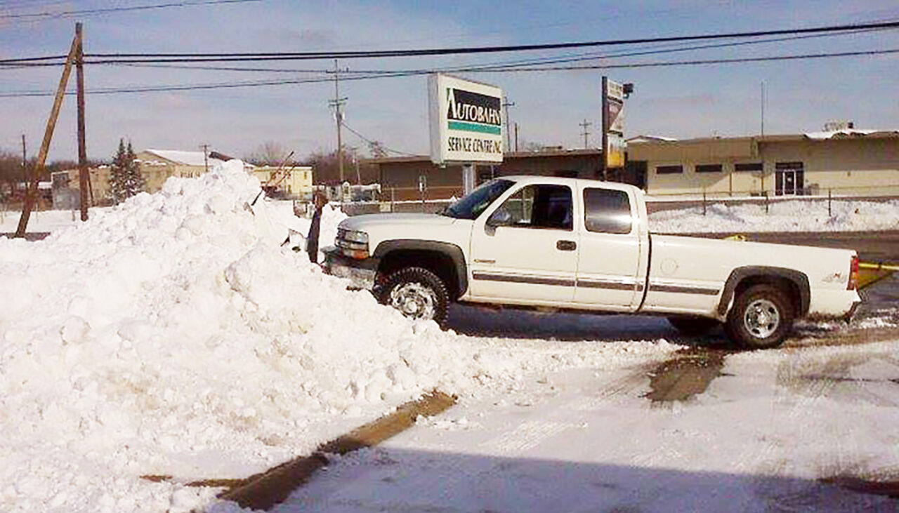 A white pickup truck is stuck with its front end buried in a large pile of snow in a parking lot near a sign that reads AUTOBARN. Snow covers the ground and the sky is clear.