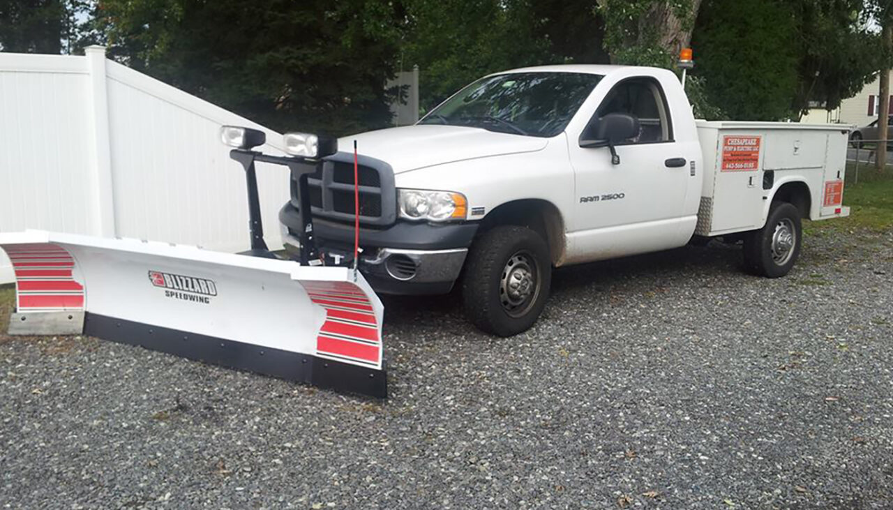 A white Ram 2500 utility pickup truck is parked on gravel, equipped with a large Bullard snow plow attached to the front. Trees and a white fence are visible in the background.