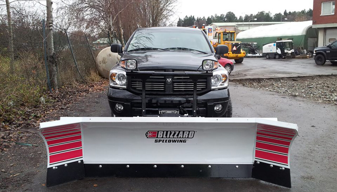 A black pickup truck is parked outdoors with a large white and red snow plow attachment labeled Blizzard Speedwing mounted on its front. Construction vehicles and buildings are visible in the background.