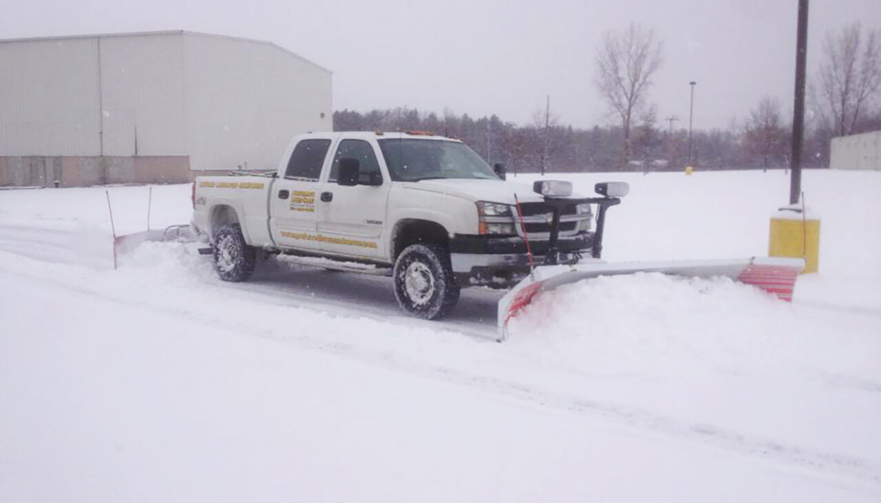 A white pickup truck equipped with a snowplow clears snow from a parking lot on a snowy day. There are buildings, trees, and snow-covered ground in the background.