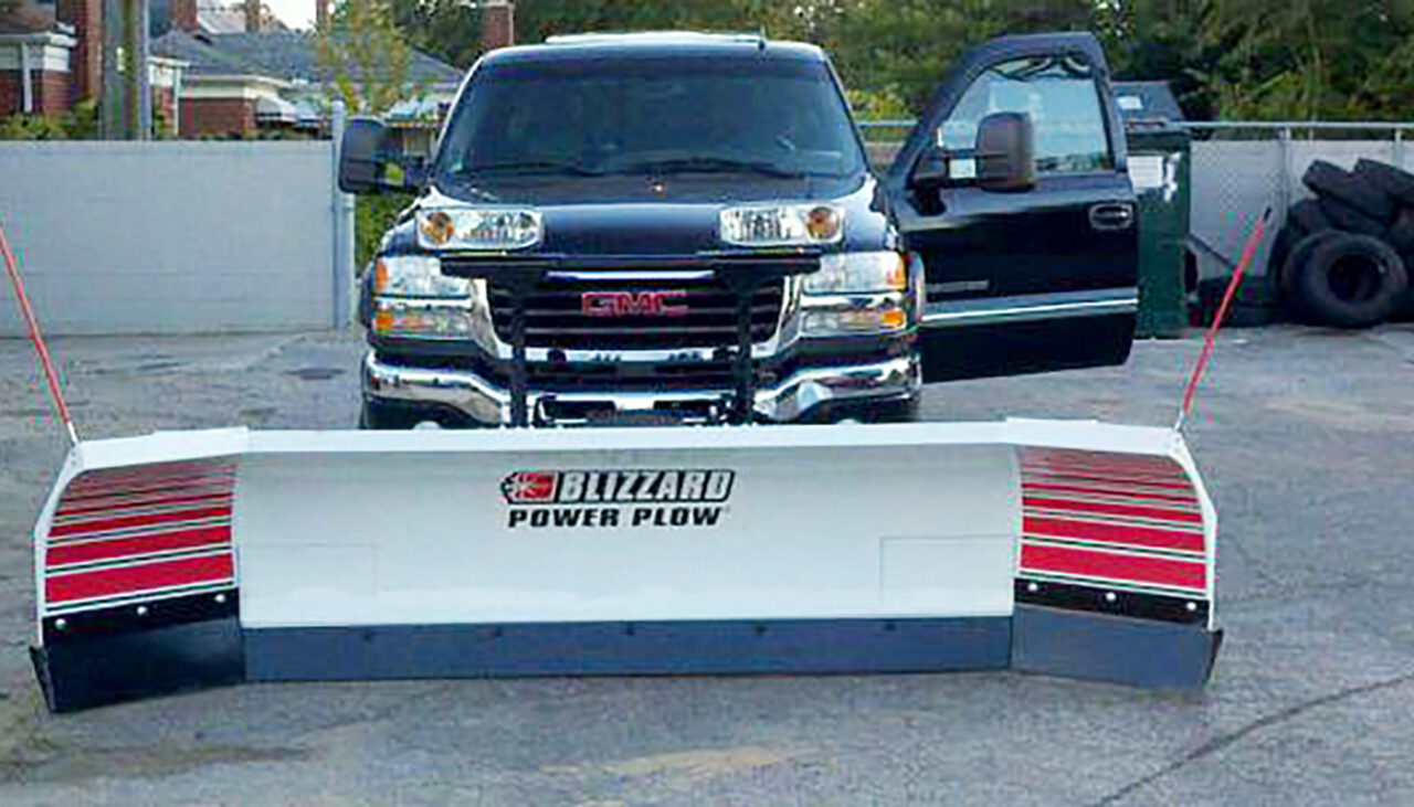 A black GMC pickup truck with its driver door open, fitted with a large Blizzard Power Plow snow plow attachment at the front, is parked on a paved surface near a fence and tires.
