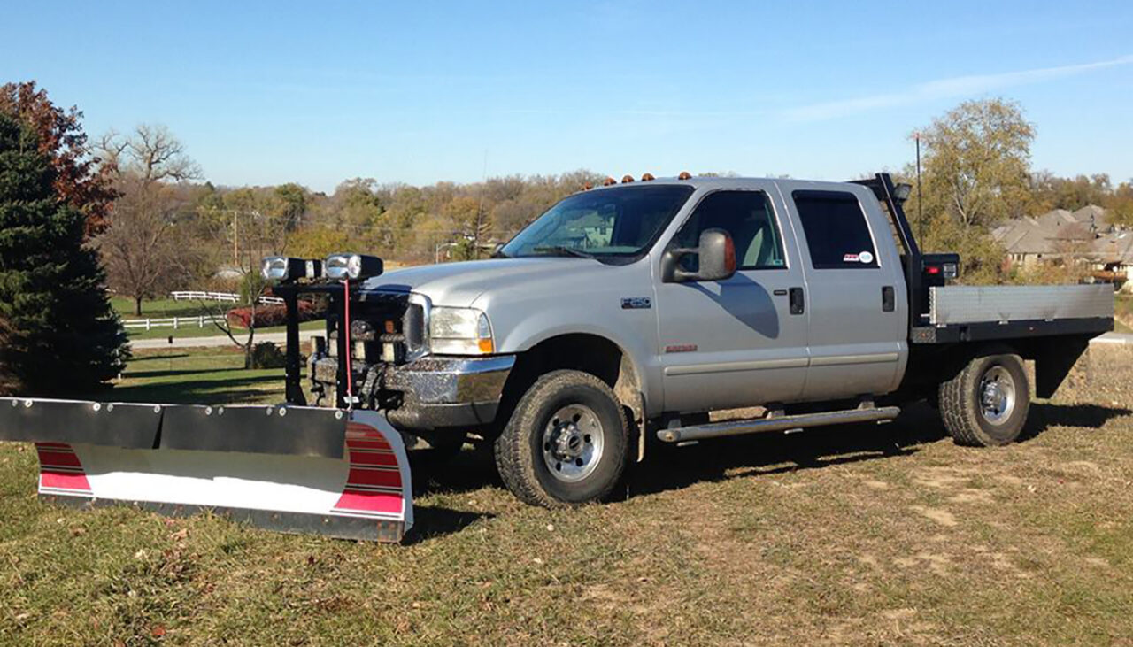 A silver pickup truck with a large snow plow attached to the front is parked on grass, with trees and houses visible in the background.