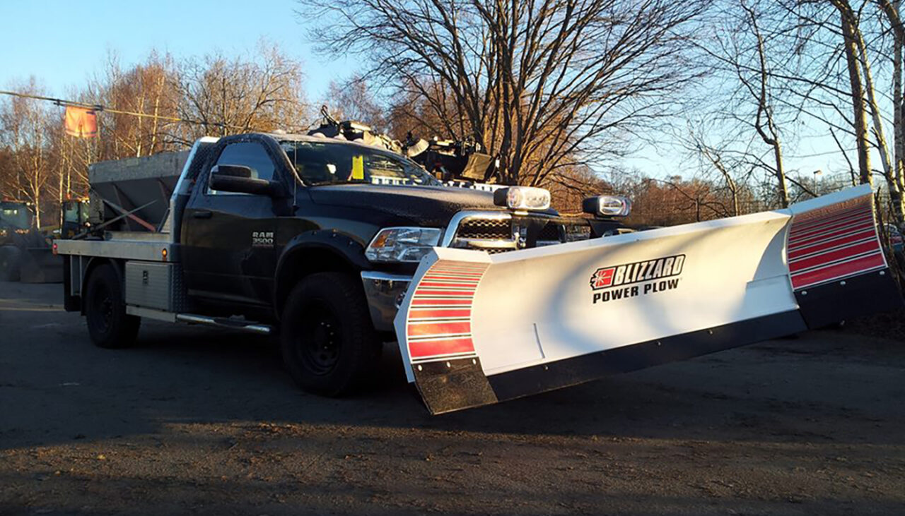 A black pickup truck equipped with a large Blizzard Power Plow snowplow attachment is parked outdoors on a paved surface, with leafless trees and a clear sky in the background.