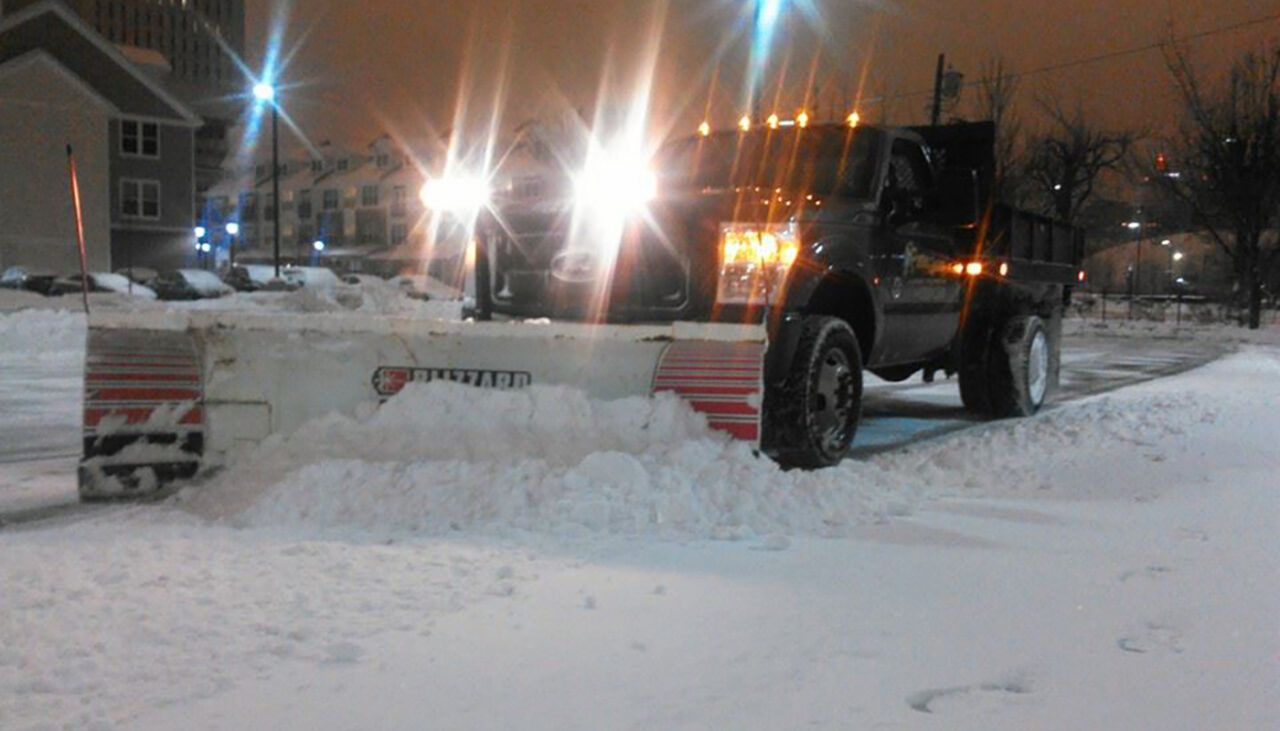 A snowplow truck with headlights on clears snow from a parking lot at night, with buildings and trees visible in the background under an orange-lit sky.