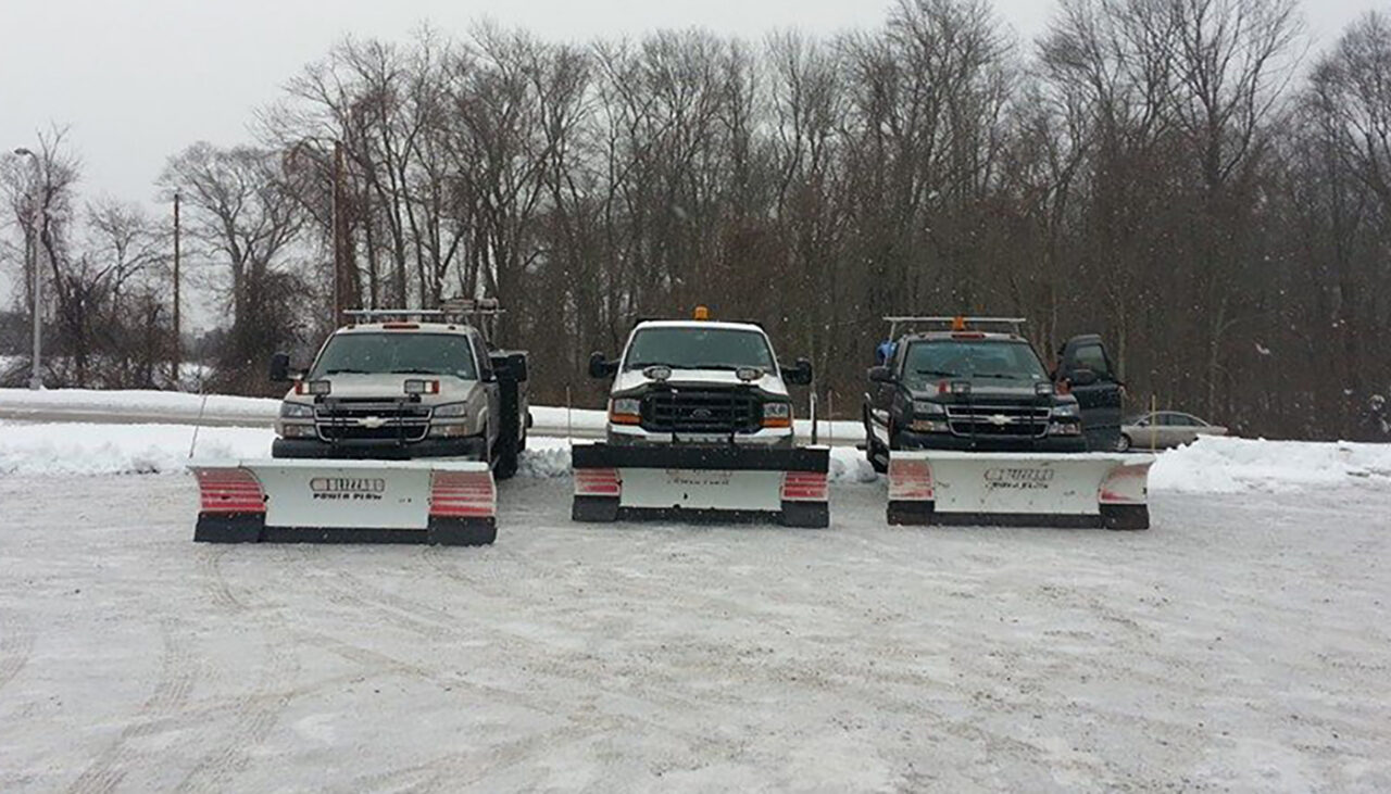 Three pickup trucks equipped with large snow plows are parked side by side on a snow-covered lot, with bare trees and a snowy landscape in the background.