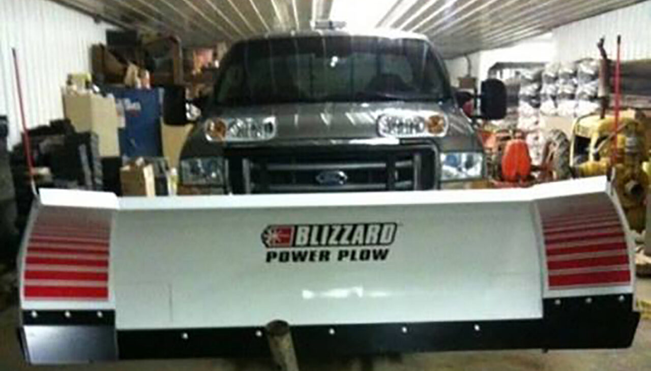 A large truck with a Blizzard Power Plow snowplow attachment is parked inside a cluttered garage with shelves, boxes, and various equipment in the background.