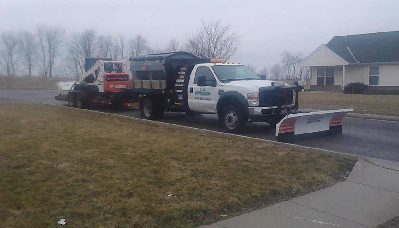A white pickup truck with a snow plow attachment and landscaping company signage is towing a trailer carrying a Bobcat loader. The scene is set on a cloudy day in a suburban neighborhood with grass and bare trees.