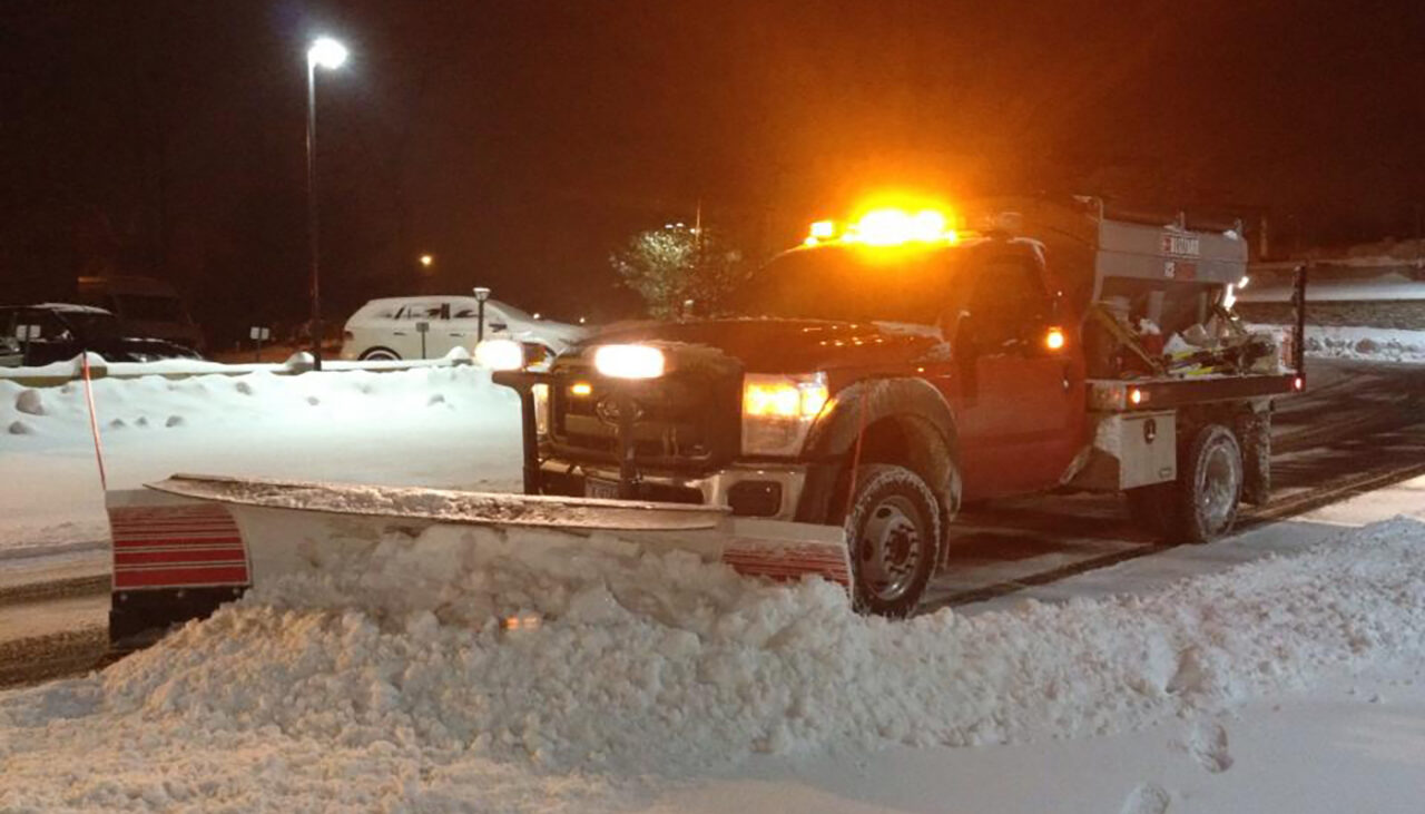 A red pickup truck with a large snow plow attached clears snow from a parking lot at night. The trucks orange warning lights are flashing, and snow is piled in front of the plow blade.