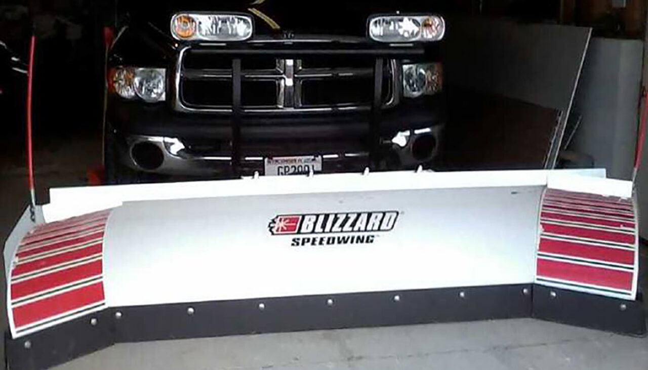 A black pickup truck with a large white Blizzard Speedwing snow plow attached to the front, parked inside a garage.