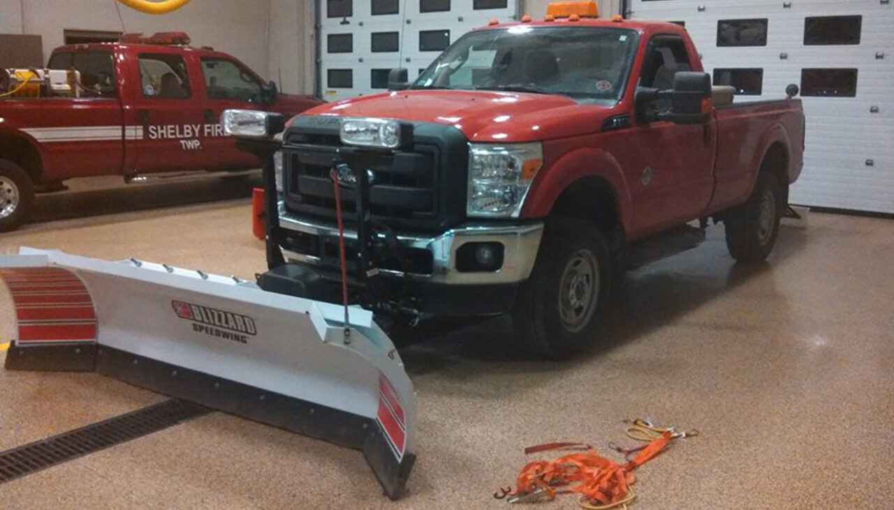 A red pickup truck equipped with a snow plow attachment is parked inside a garage. A coiled orange tow strap lies on the floor nearby, and another red emergency vehicle is visible in the background.