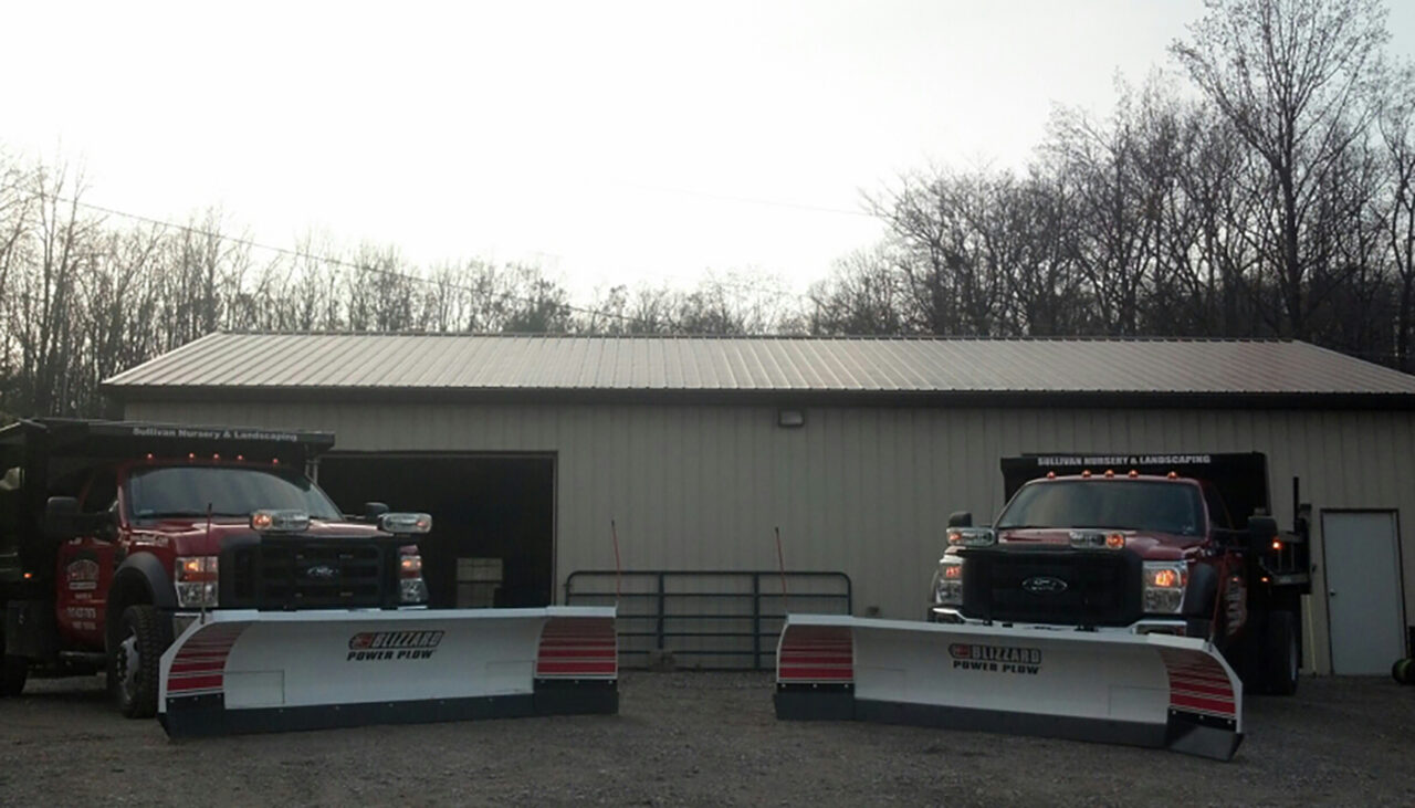 Two red trucks with attached snow plows are parked in front of a metal building with two large garage doors. Bare trees are visible in the background under a bright, hazy sky.