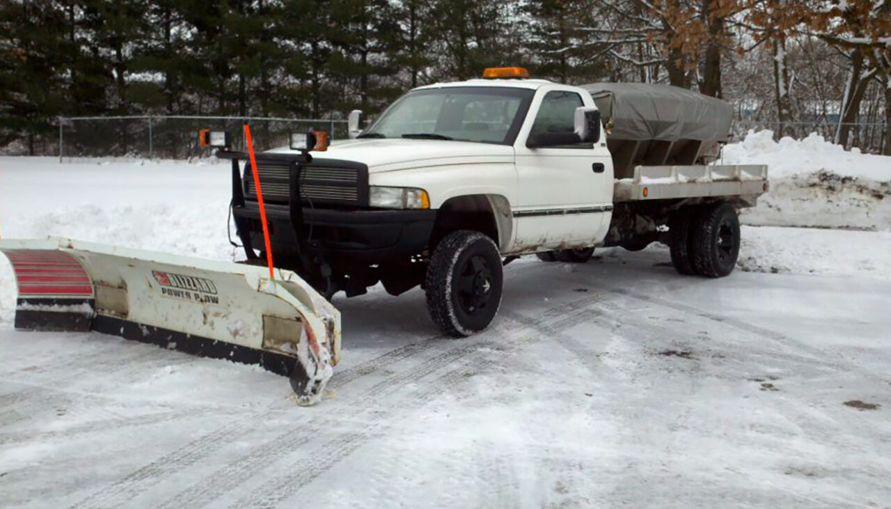A white pickup truck with a snow plow attachment is parked on a snowy surface, with snow piled and trees in the background. The truck has an amber warning light on top and a covered load in the bed.