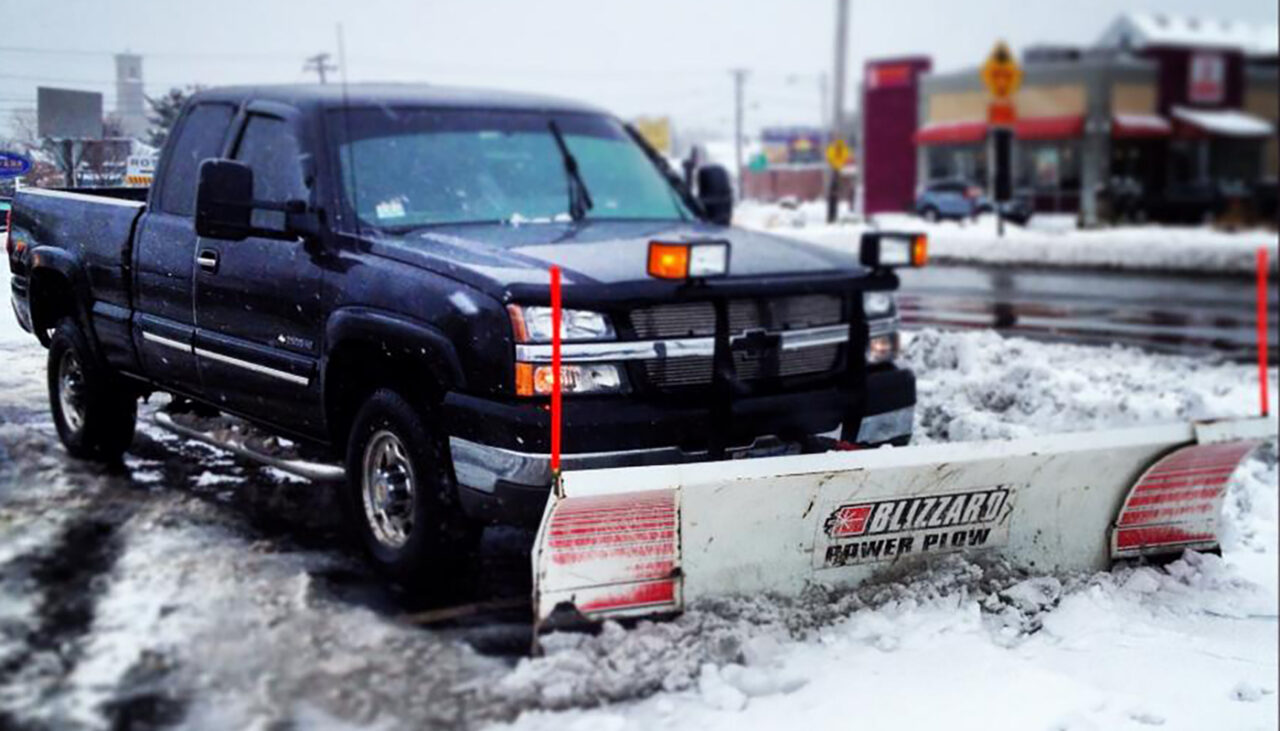 A black pickup truck equipped with a Blizzard Power Plow snow plow attachment is parked in a snowy parking lot, with snow covering the ground and buildings visible in the background.
