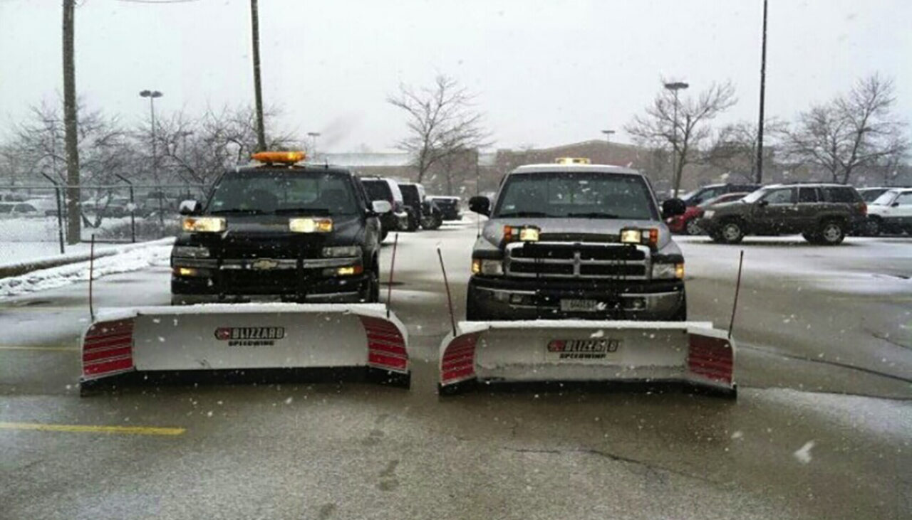 Two black pickup trucks equipped with large snow plows are parked side by side in a snowy parking lot. Snow is falling, and several cars and leafless trees are visible in the background.