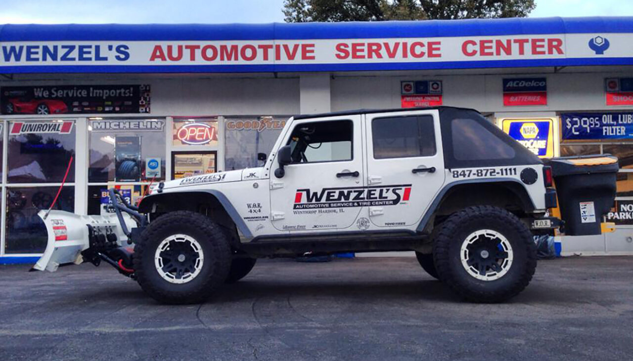 A white Jeep Wrangler with off-road tires and a front snow plow attachment is parked in front of Wenzels Automotive Service Center. The storefront has various tire brand signs and large windows.