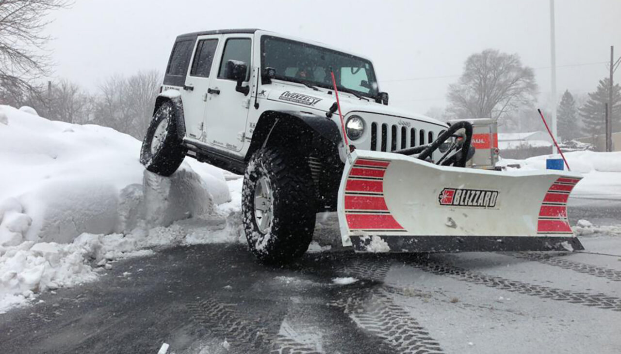 A white Jeep with a large snow plow attachment clears snow in a parking lot, with one front wheel lifted onto a pile of snow. Snow covers the ground and trees in the background.
