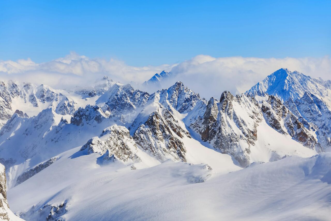 Snow-covered mountain peaks under a clear blue sky, with some clouds settling around the higher summits. The rugged terrain is highlighted by bright sunlight and sharp shadows.
